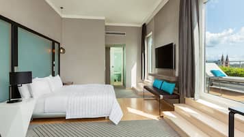 Ein modernes Hotelzimmer mit einem großen Bett, einem Fernseher, einem Balkon mit Blick und einer Sitzecke mit Kissen.