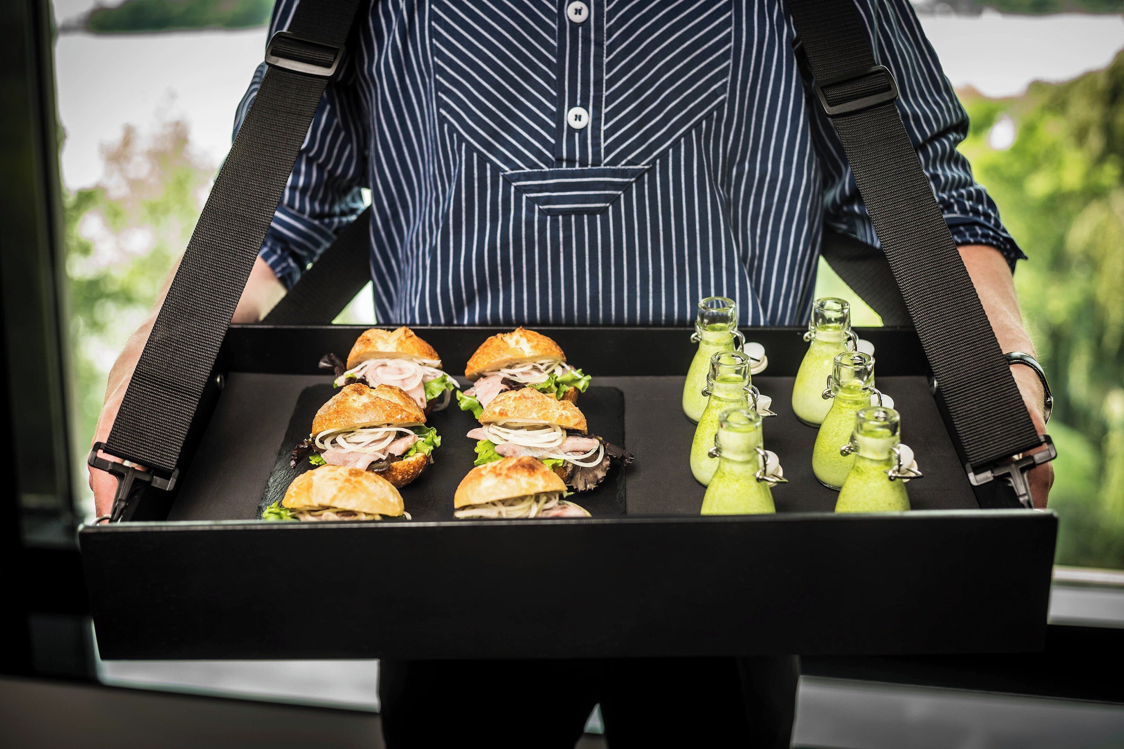 Eine Person hält ein Tablett mit Sandwiches und grünen Getränken.