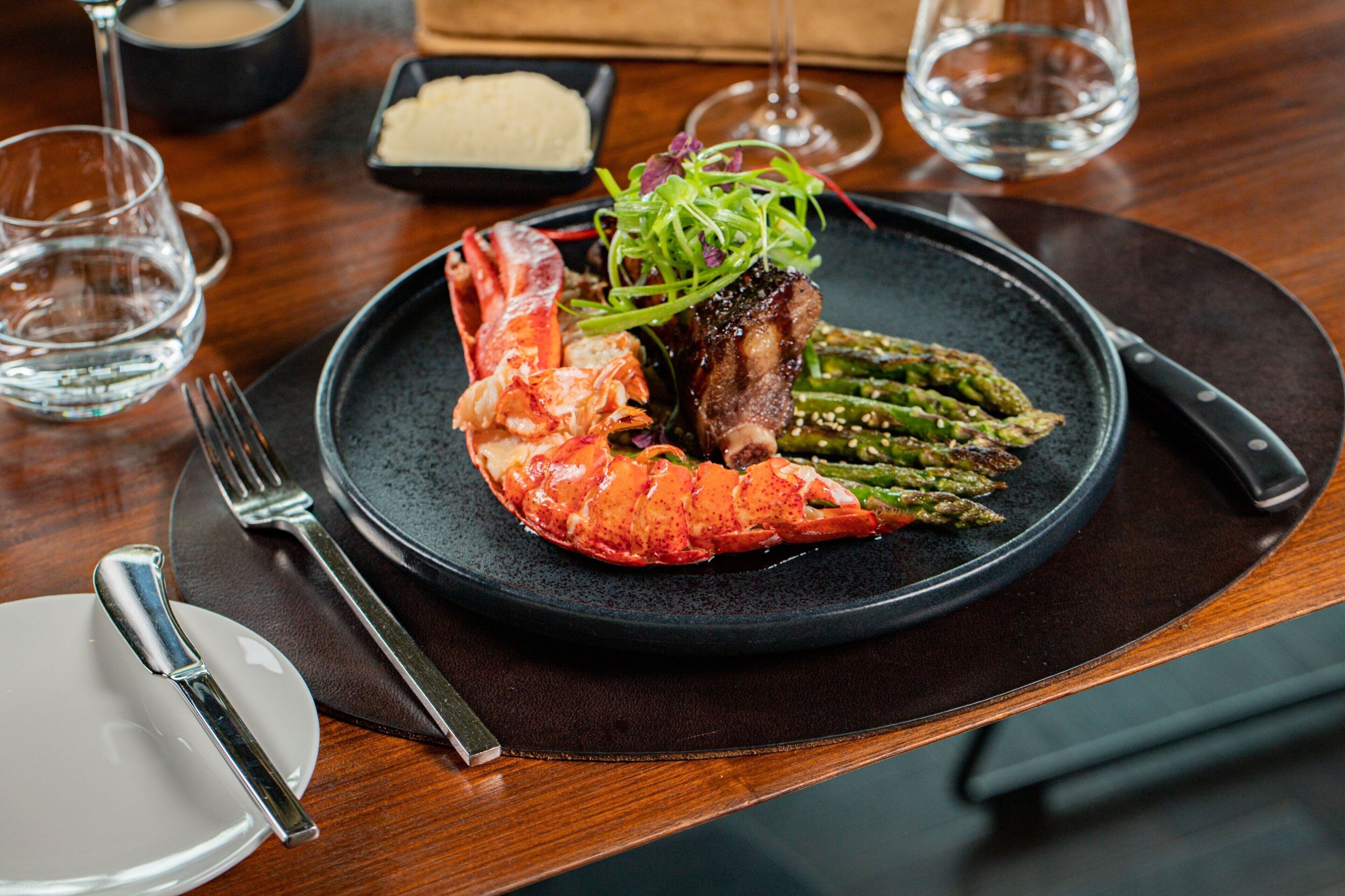 A plate of lobster and asparagus on a dark plate, with a fork and knife on the left.