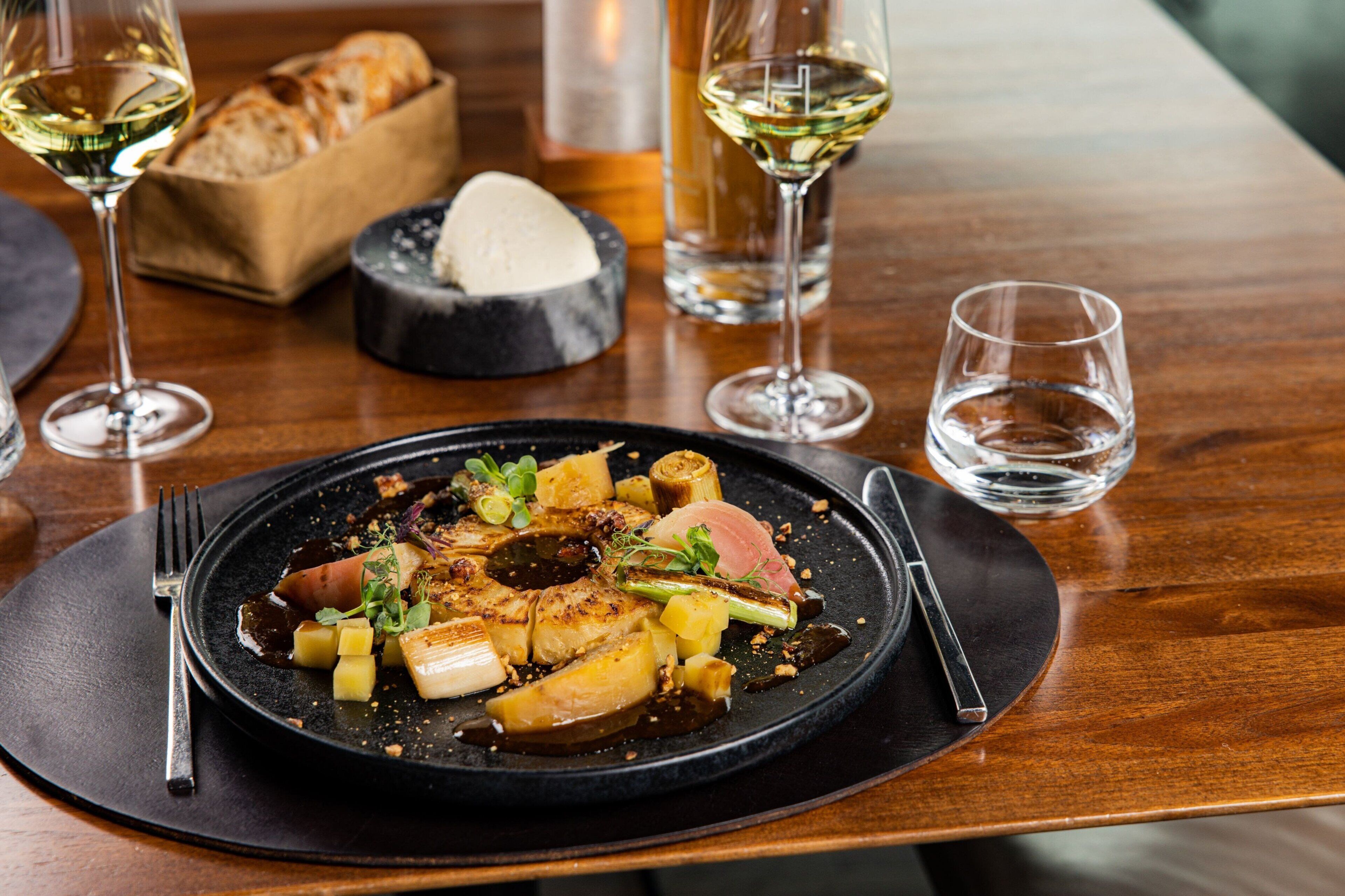 A dining table with a black plate of food, two wine glasses, and a bread basket.