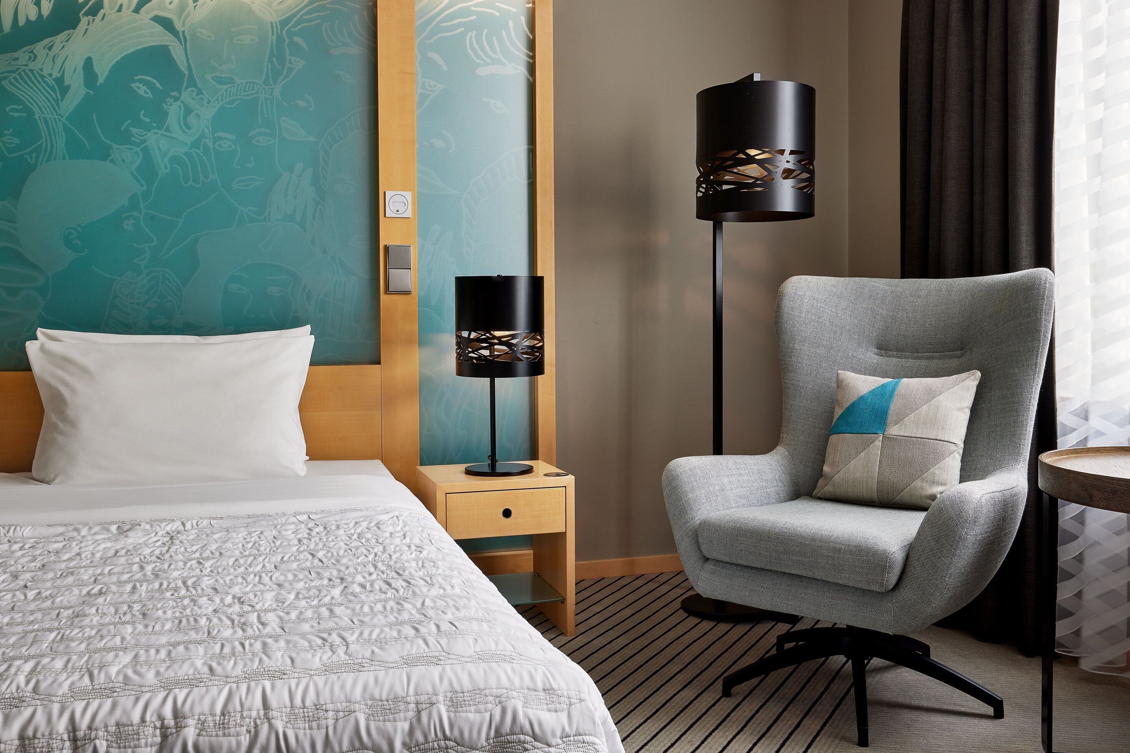 A hotel room with a bed, a grey armchair, a standing lamp, and a wall with a mural.
