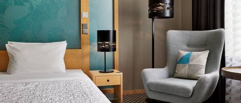 A hotel room with a bed, a grey armchair, a standing lamp, and a wall with a mural.