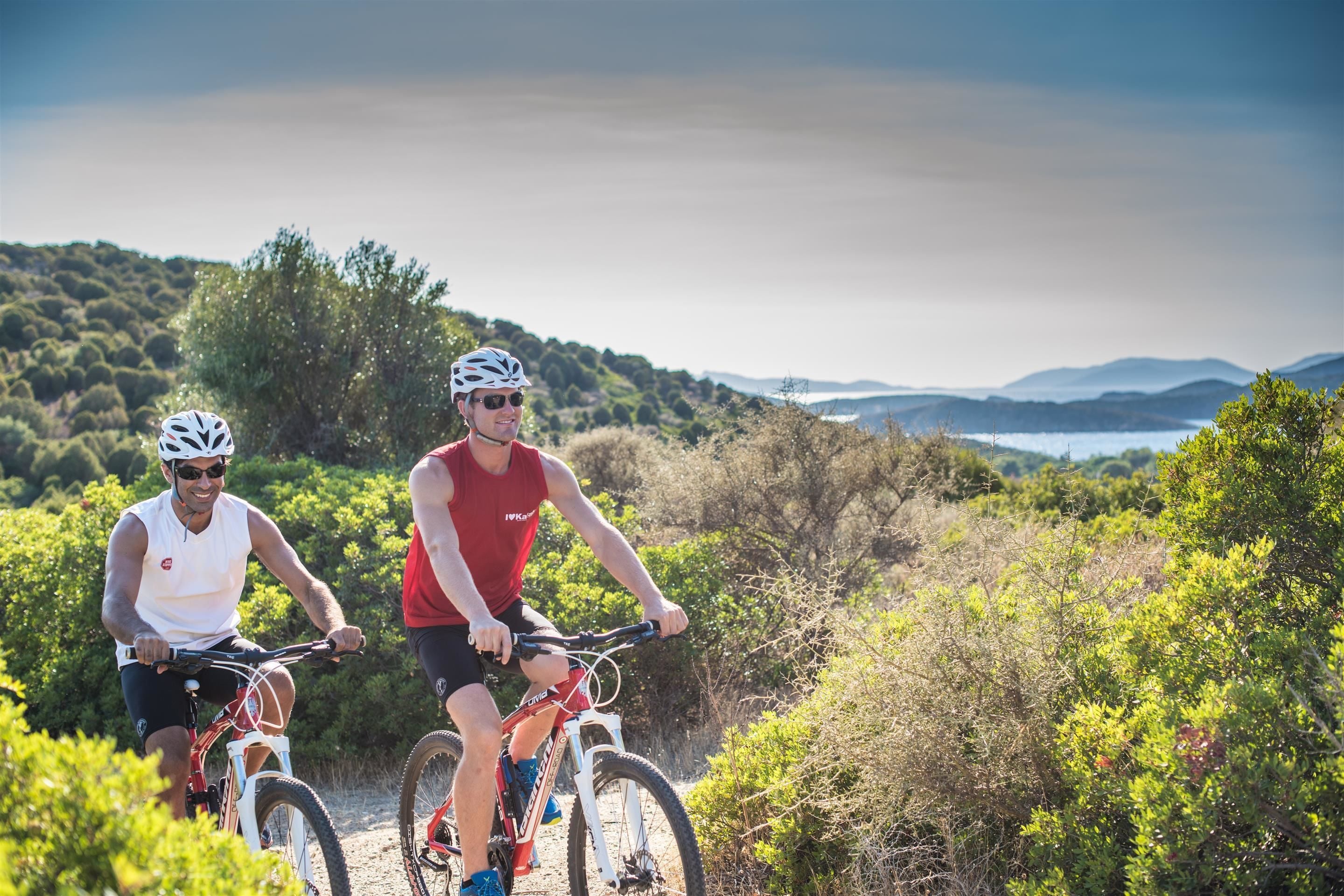 Two cyclists on a trail with a scenic view of hills and water in the background.