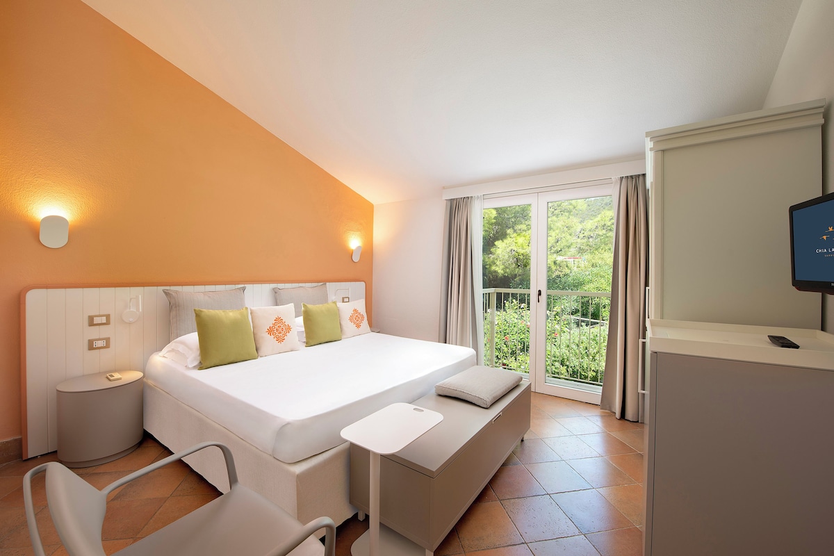 Une chambre d’hôtel avec un lit, des tables de chevet, une télévision, un banc et une vue sur la verdure par la fenêtre.