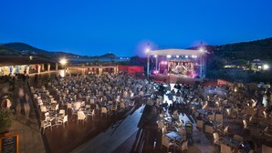 An outdoor dining area with tables and chairs, a stage with lighting, and a mountainous backdrop.