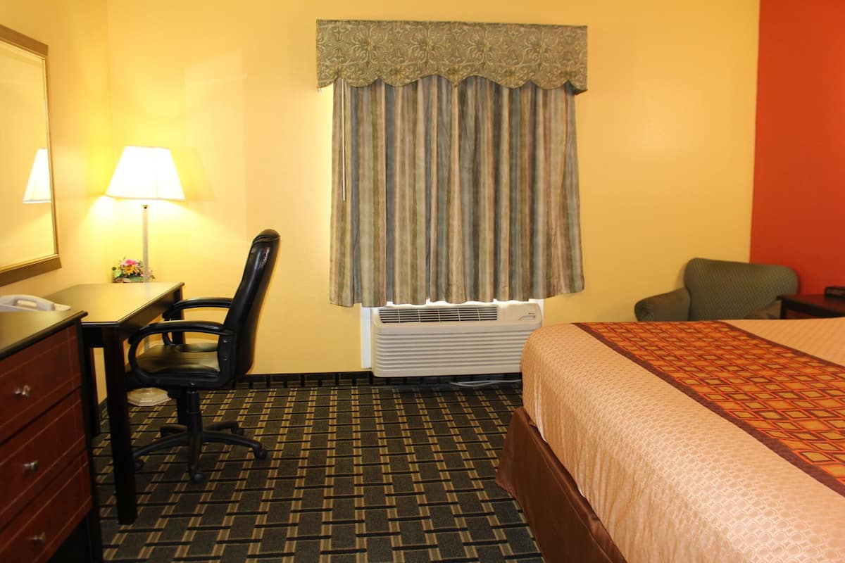 A hotel room with a bed, a desk with a lamp, a chair, an air conditioner, and a window with curtains.