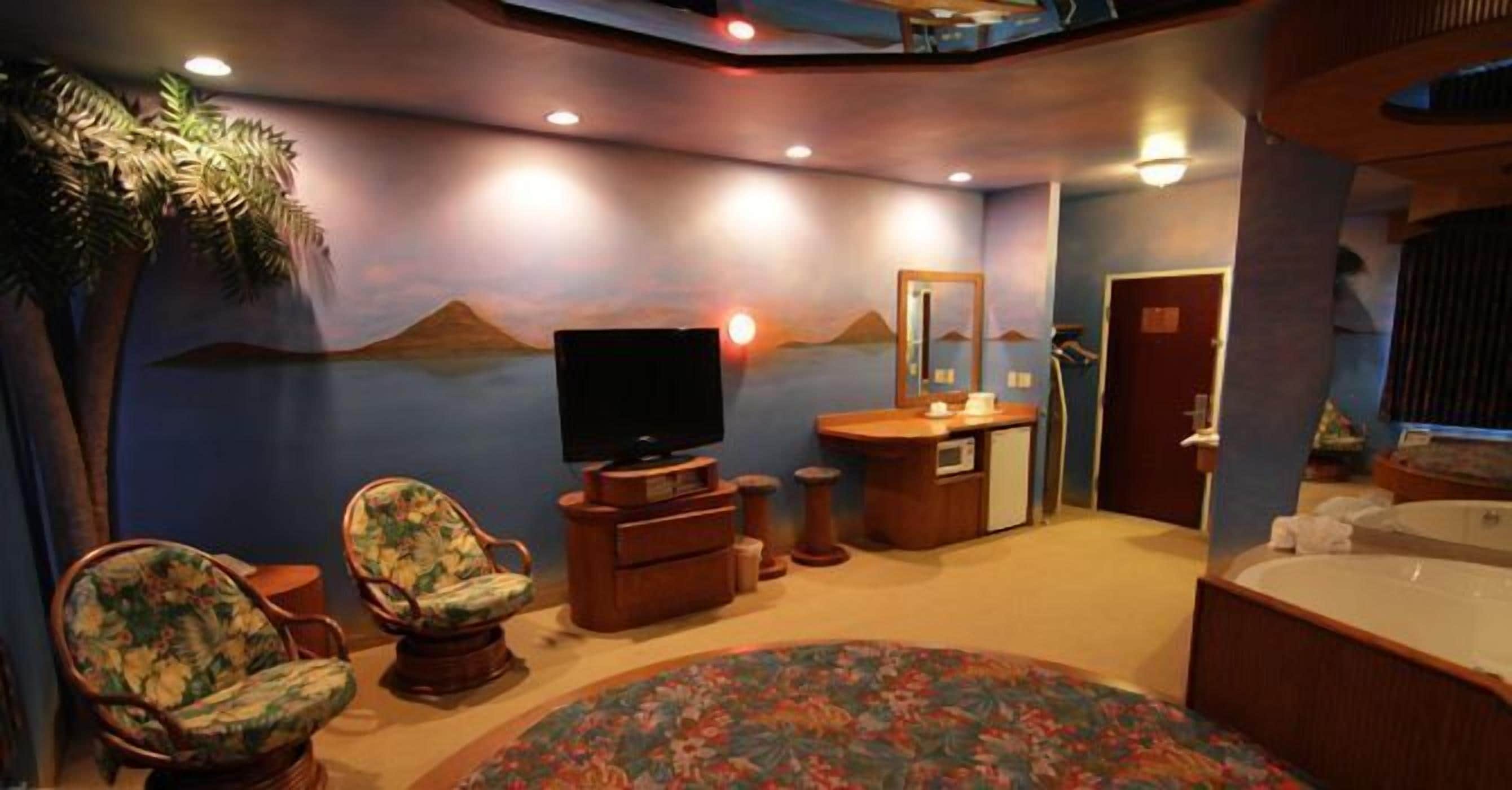 Ein Zimmer mit Fernseher, zwei Sesseln, einem kleinen Schreibtisch, einem Spiegel und einer Badewanne.