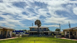Una casa de dos pisos con balcón, rodeada por un cerco de piquetes blancos, bajo un cielo azul despejado con algunas nubes.