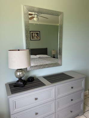 Un dormitorio con cama, mesita de noche con lámpara, cómoda con cajones, espejo y ventilador de techo.