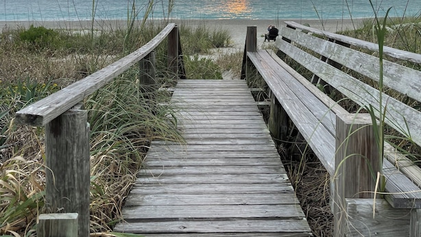 Una pasarela de madera que conduce a un banco con vista a una playa y un atardecer en la distancia.