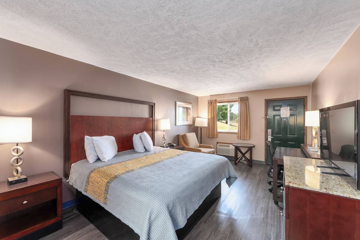 A hotel room with a large bed, bedside tables, a desk, a chair, a TV, and a door.