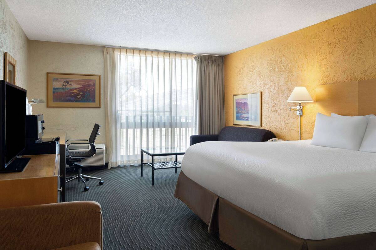 A hotel room with a large bed, a desk with a computer, a chair, a desk lamp, and a window with curtains.