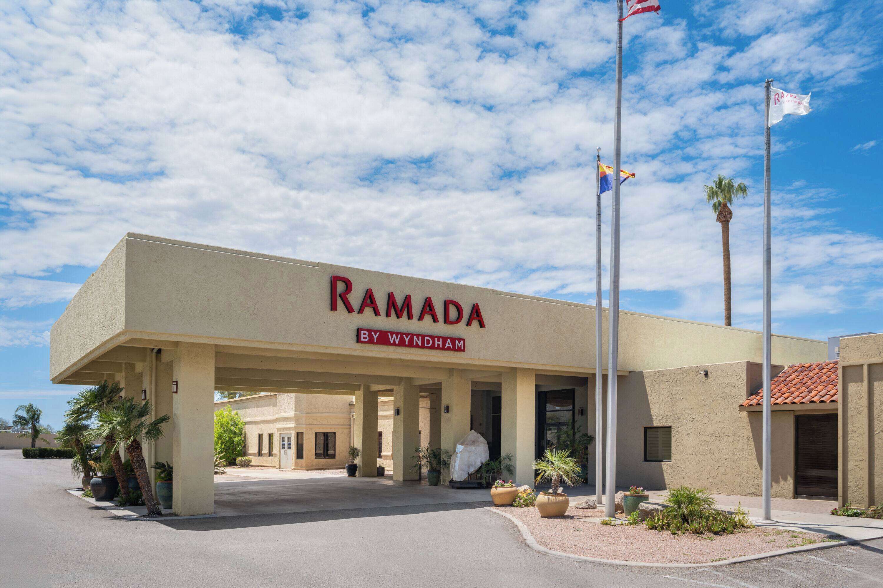 Un hôtel appelé Ramada by Wyndham, doté d’une entrée couverte, de palmiers et de drapeaux.