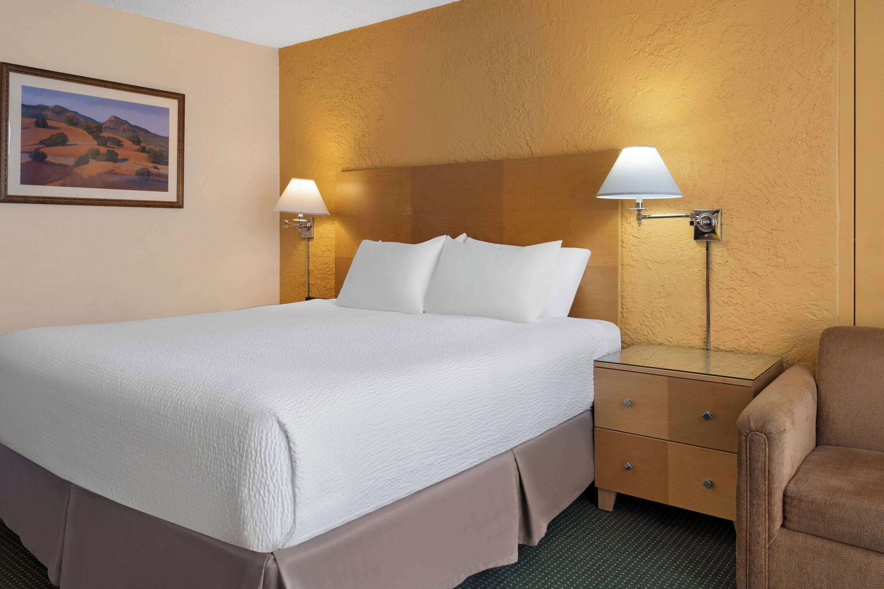 A hotel room with a large bed, two bedside lamps, a wooden headboard, a small wooden nightstand, and a brown armchair.