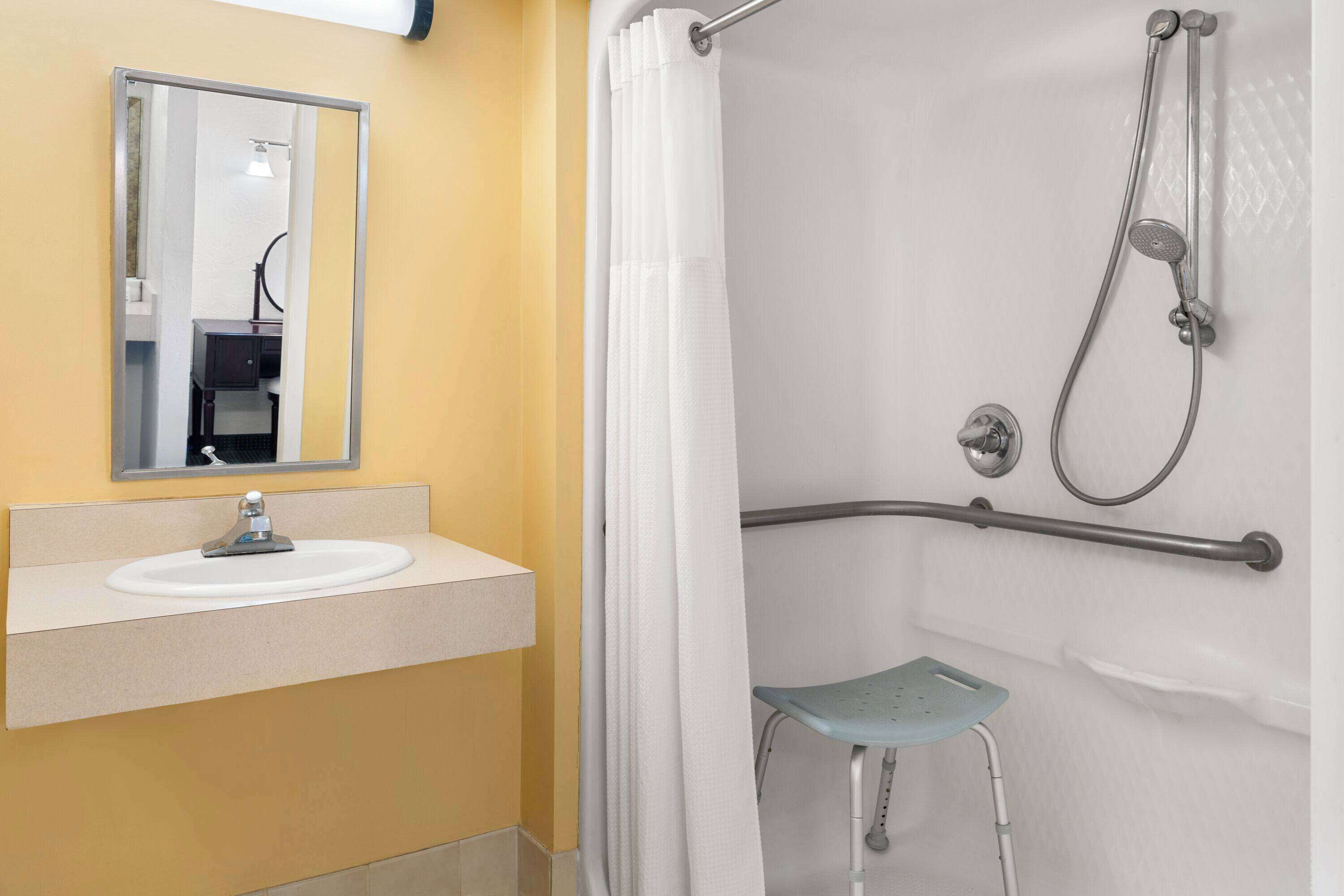 Un baño con lavamanos, espejo, ducha y un banco accesible para silla de ruedas.