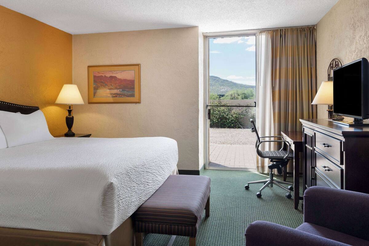 A hotel room with a bed, a desk with a television, a chair, and a window with a view of the outdoors.
