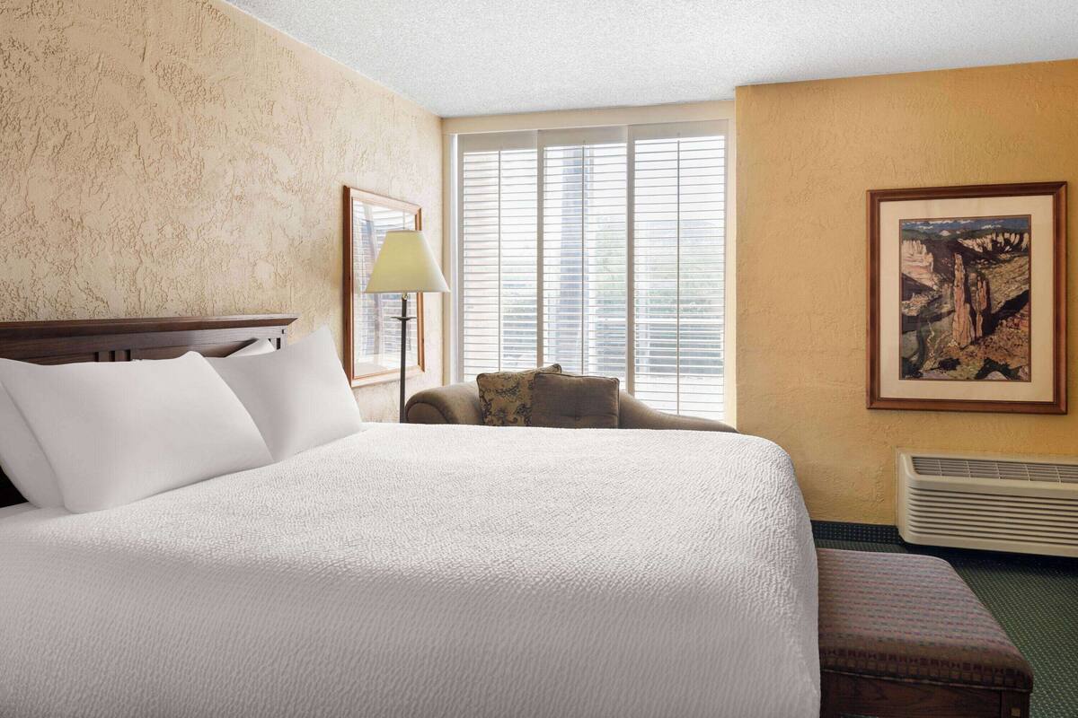 A hotel room with a large bed, a chair, a lamp, a framed picture, and a window with blinds.