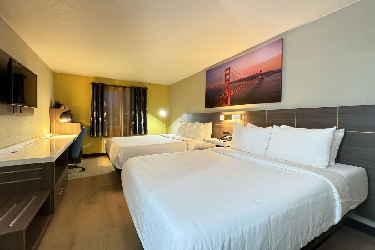 A hotel room with two beds, a desk, a chair, a TV, and a painting of a bridge.