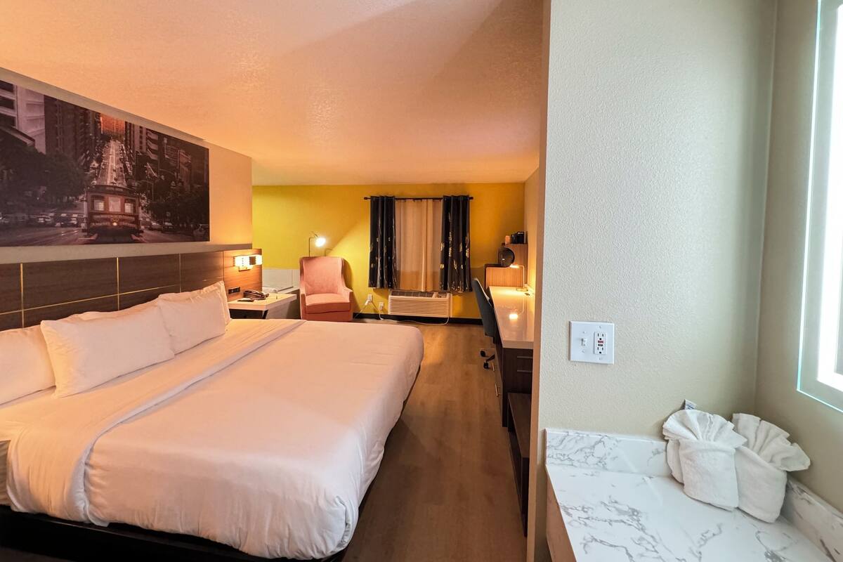 A hotel room with a large bed, a desk with a chair, a small table with a lamp, and a bathroom with a marble countertop.