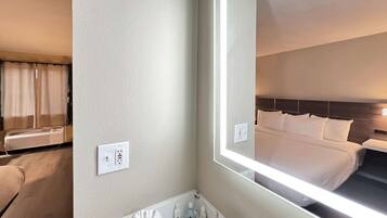 A hotel room with a large mirror reflecting a neatly made bed, a bathroom with a marble countertop, and a wall-mounted air conditioning unit.