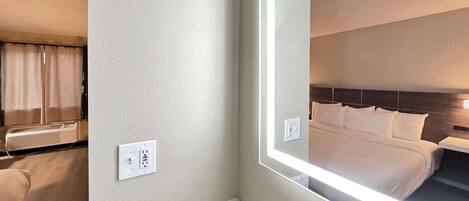 Habitación de hotel con un espejo grande que refleja una cama bien hecha, un baño con encimera de mármol y un equipo de aire acondicionado de pared.