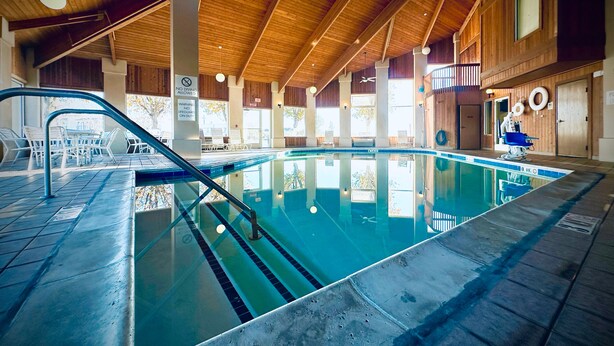 Una piscina interior con techo de madera, paredes de vidrio transparente y una silla para socorrista.