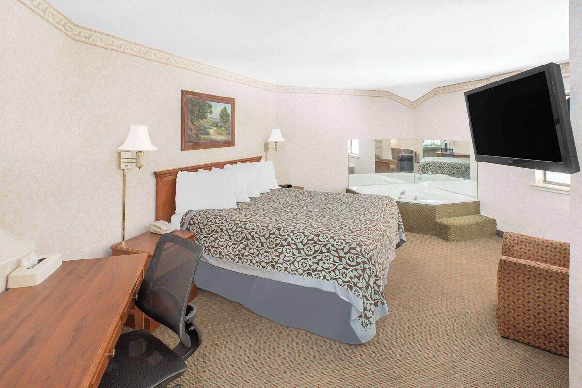 A hotel room with a large bed, a desk, a chair, a TV, a bathroom with a bathtub, and a mirror.
