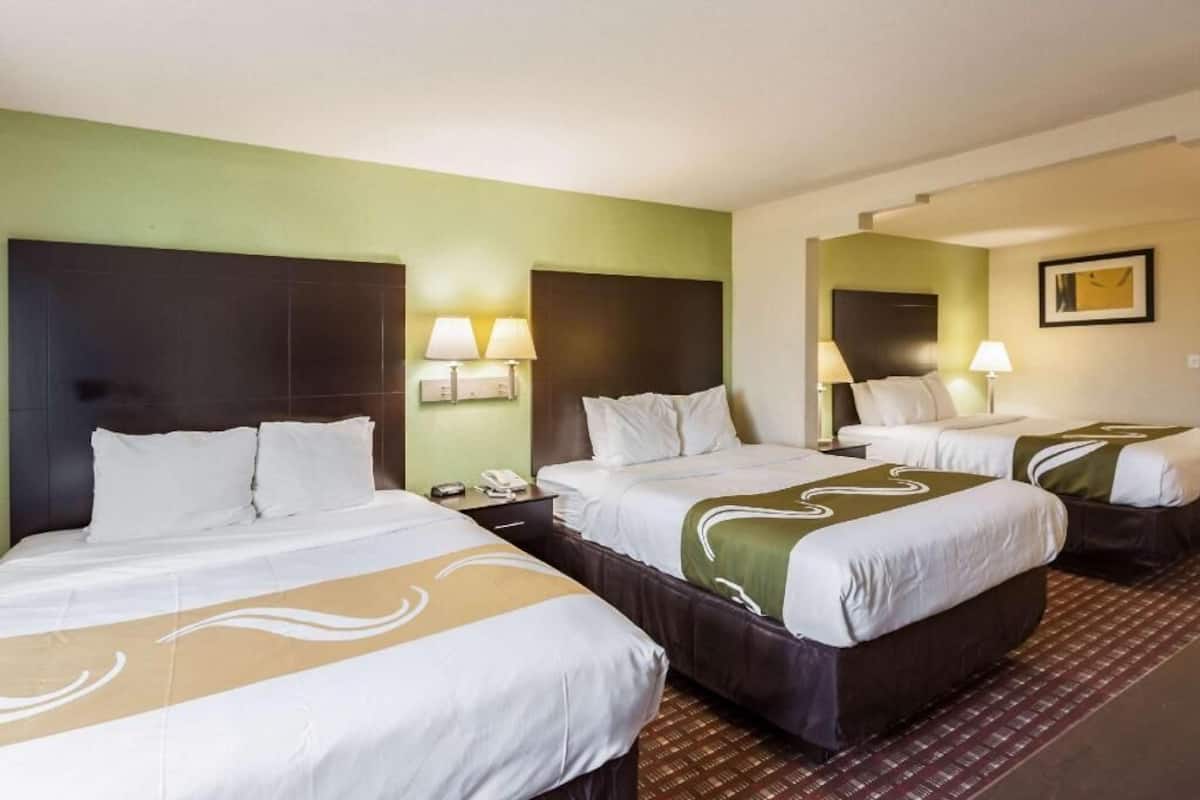 Una camera d'albergo con tre letti, ognuno con biancheria da letto bianca e un tocco di verde. Sono presenti abat-jour, un telefono sul comodino e un quadro incorniciato appeso alla parete.