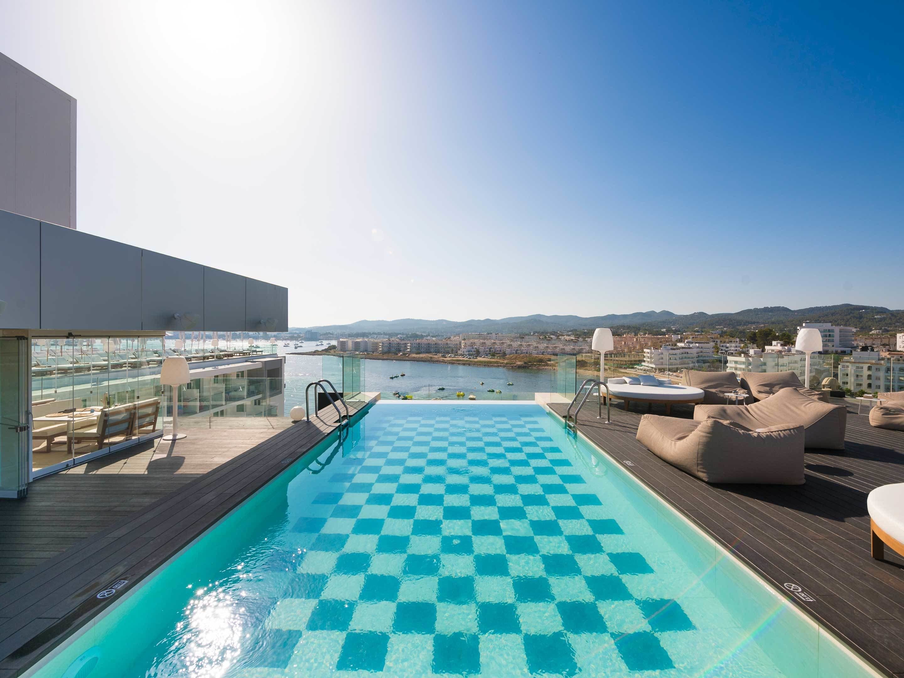 A rooftop pool with a checkered floor, lounge chairs, and a view of a marina and cityscape.