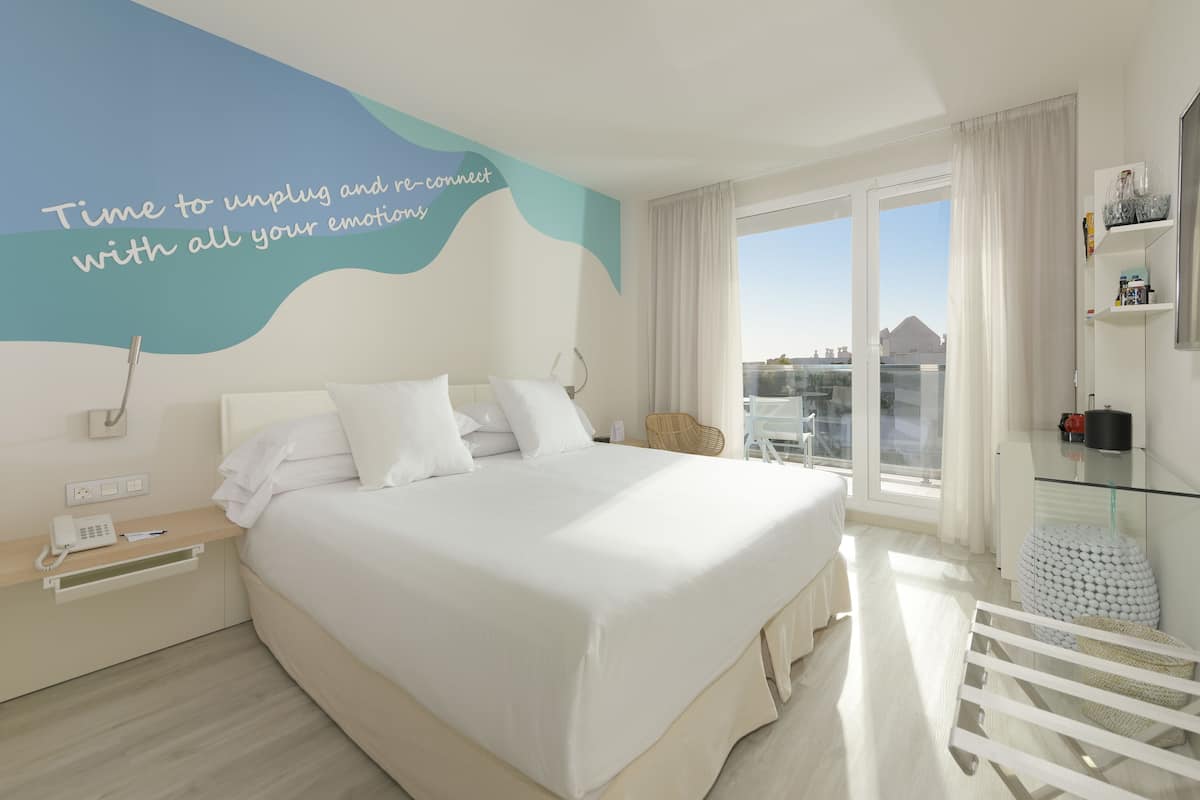 Quarto de hotel moderno com uma cama grande, mesa de cabeceira com telefone e caneta, mesa de centro de vidro, uma cadeira e vista para a cidade através de grandes janelas.