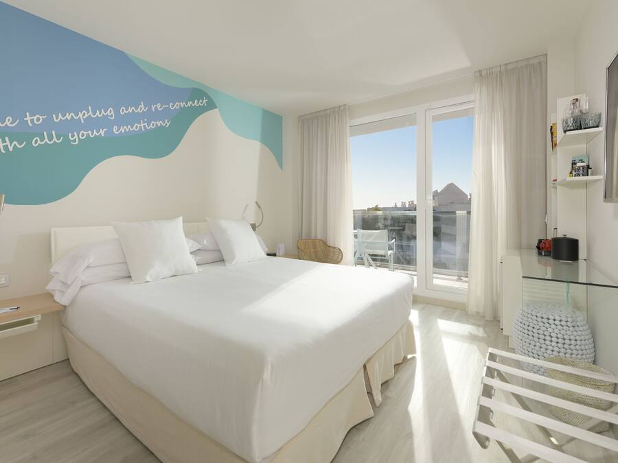 A modern hotel room with a large bed, a bedside table with a phone and a pen, a glass coffee table, a chair, and a view of the city through large windows.