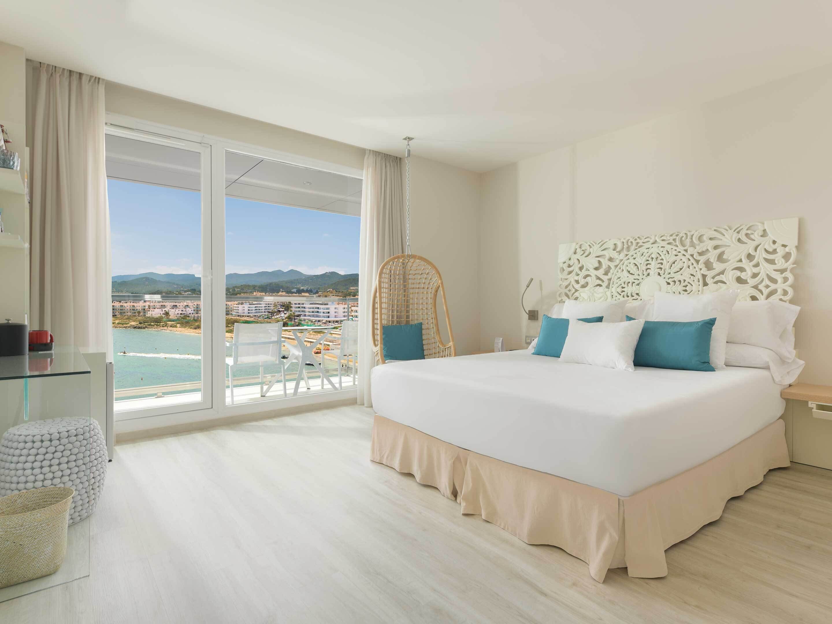 Camera d'hotel con un ampio letto, vista sulla spiaggia e sulla città, una poltrona in vimini e un tavolino in vetro.
