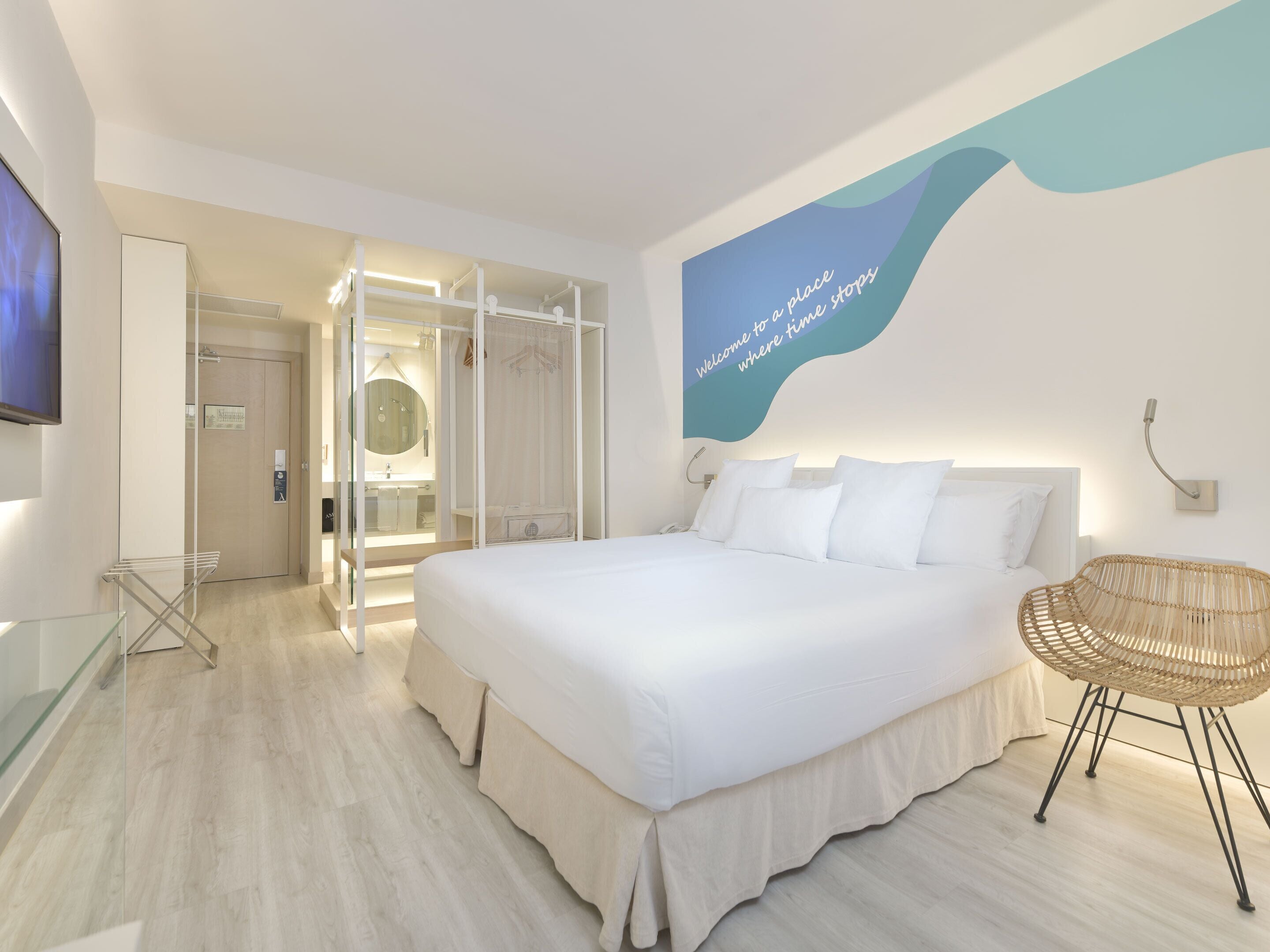 Una habitación de hotel moderna con una cama grande, un tendedero de vidrio, una silla de mimbre y un televisor montado en la pared.