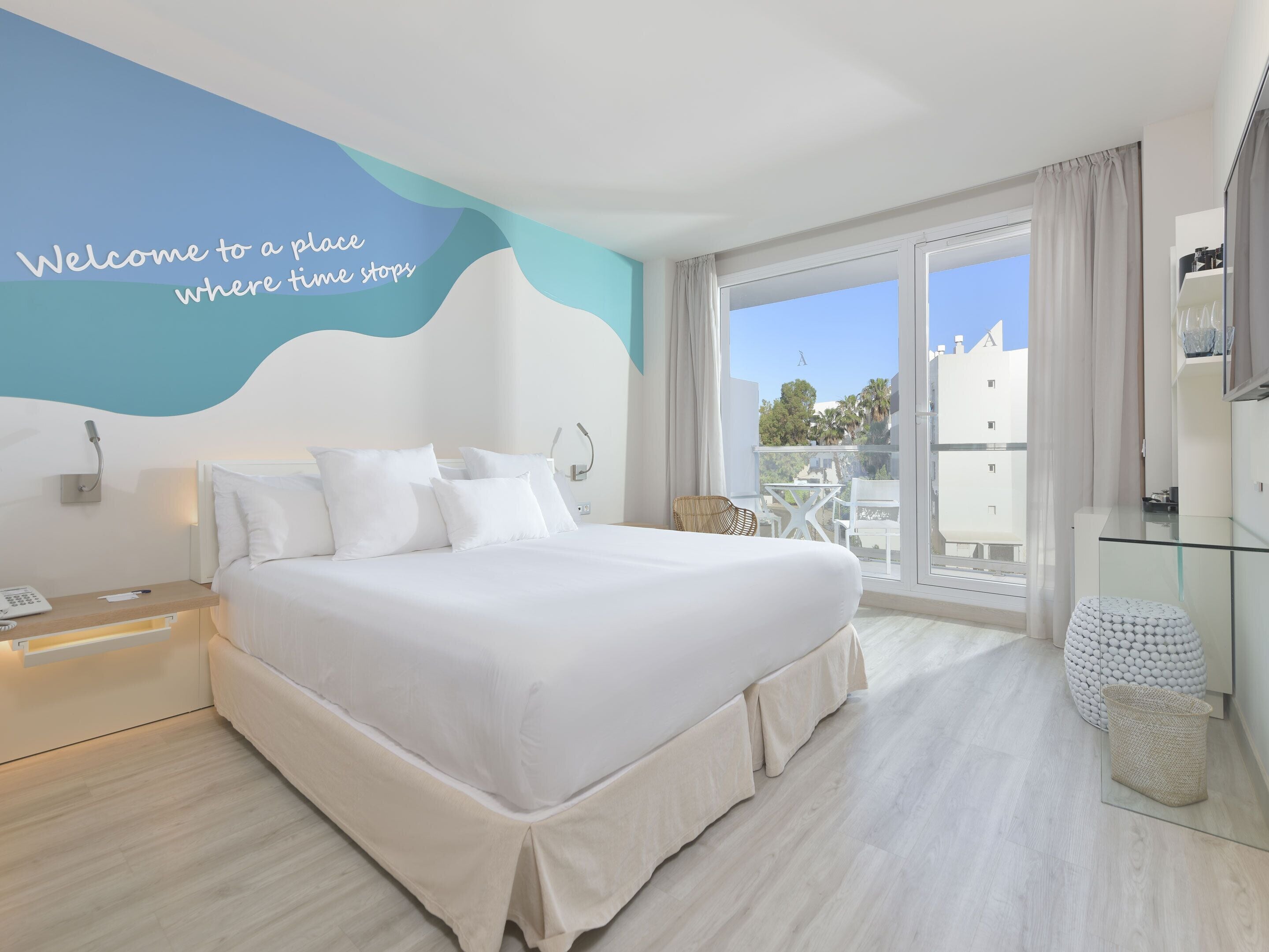 A modern hotel room with a large bed, a balcony with a view, and a wall with a welcoming message.