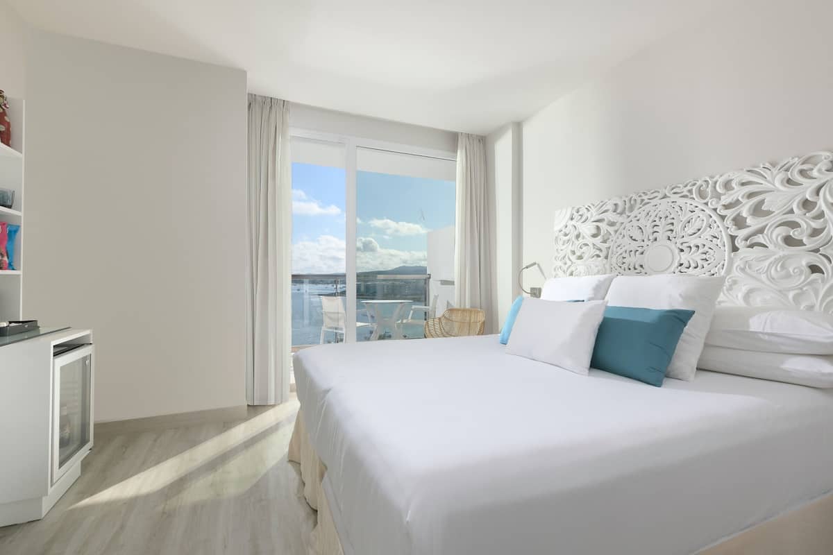 Quarto de hotel com cama grande, cabeceira branca, varanda com vista para a cidade e escrivaninha com cadeira.