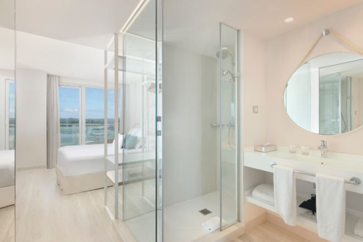 Quarto de hotel moderno com chuveiro em cabine de vidro, uma cama grande, banheiro com toucador e espelho, e vista da cidade.