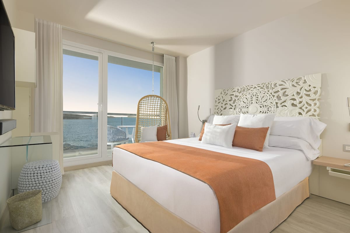 Quarto de hotel moderno com uma cama grande, uma cadeira de vime, uma mesa de vidro e vista para o mar.