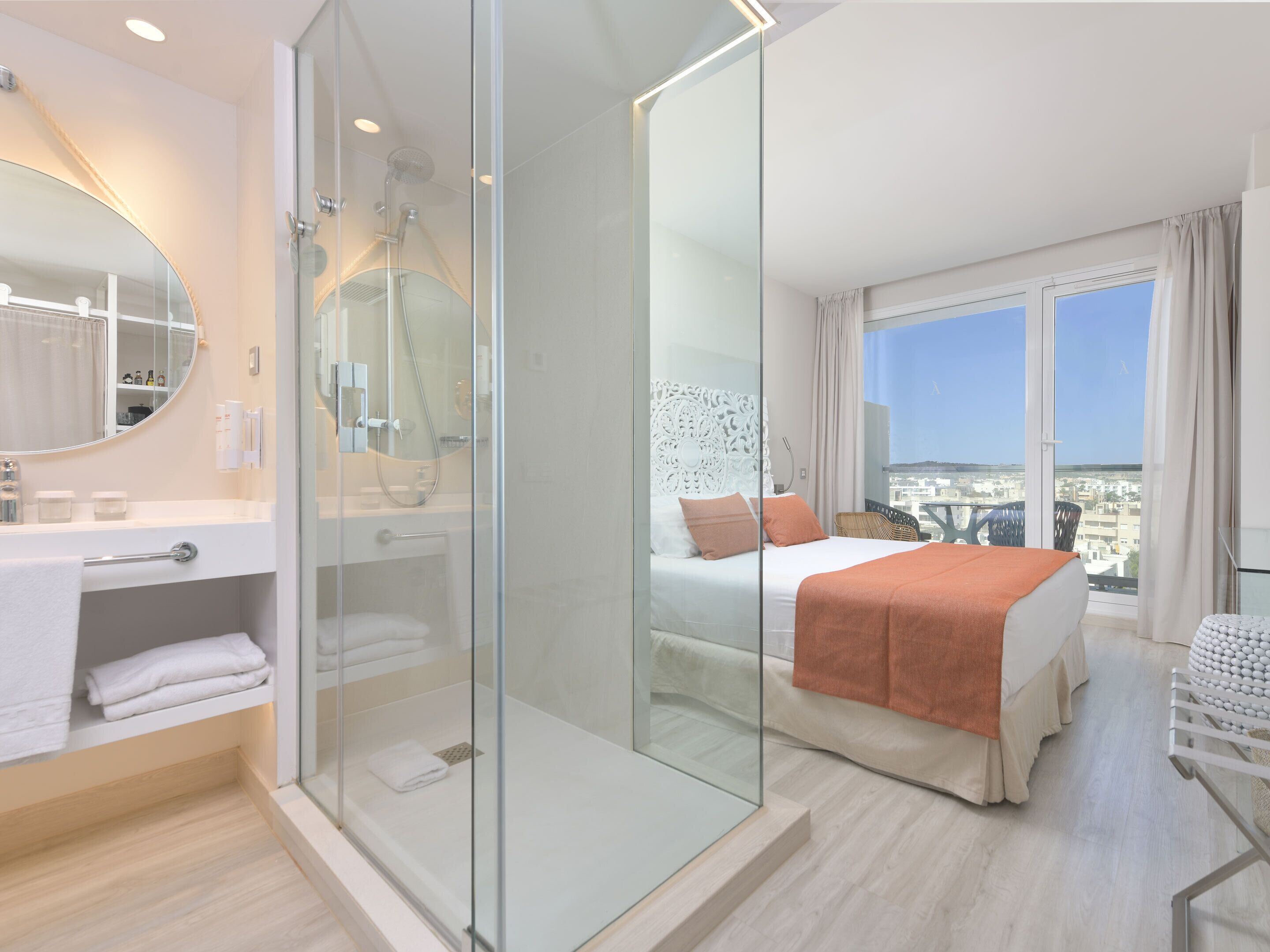 Une chambre d’hôtel moderne dotée d’une douche en verre, d’un grand lit avec un couvre-lit orange, d’une tête de lit blanche et offrant une vue sur la ville par la fenêtre.