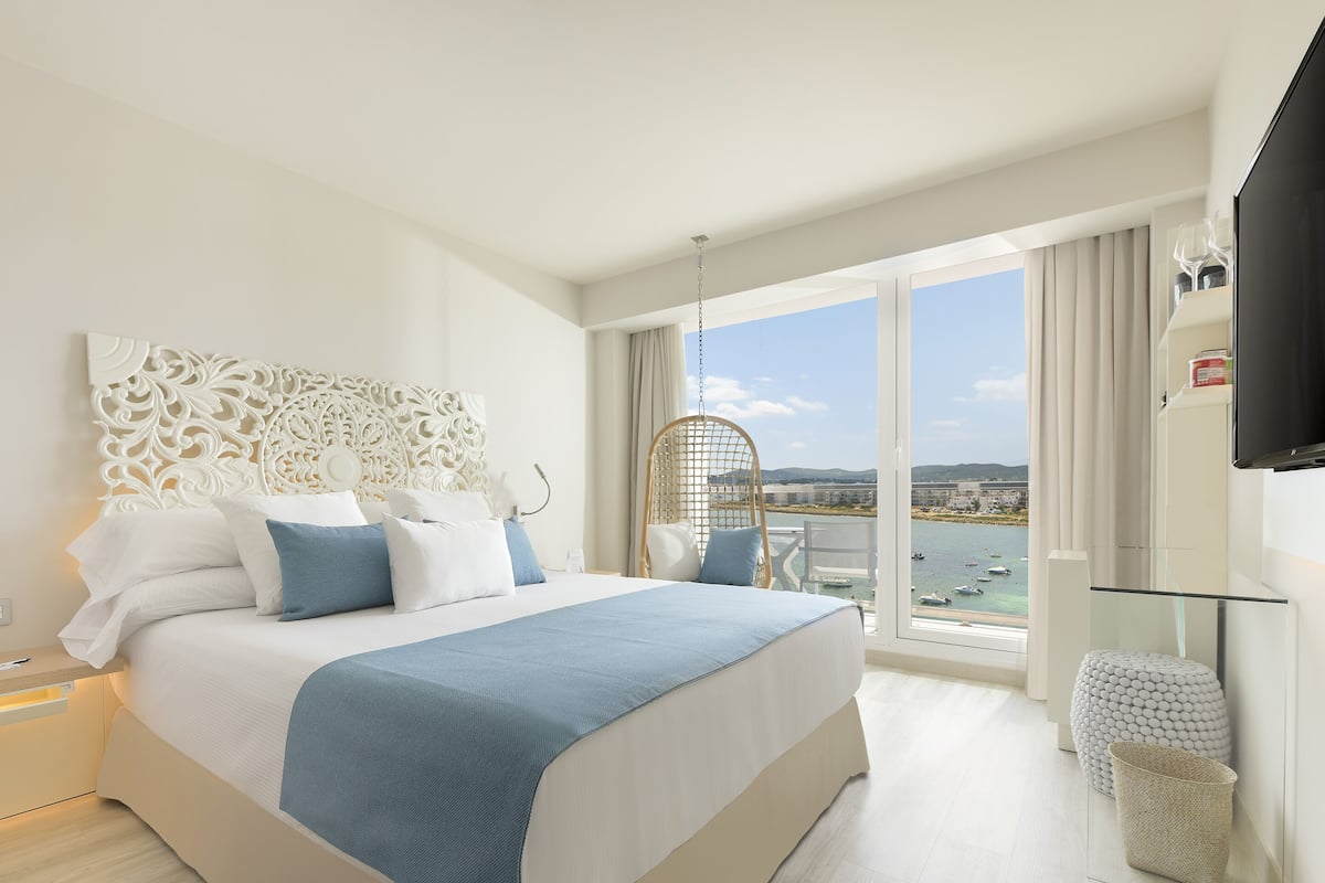 Quarto de hotel moderno com uma cama grande, cabeceira decorativa, varanda com vista para o mar e uma televisão fixada na parede.