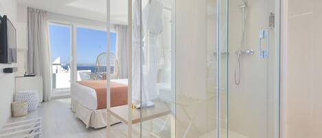 Ein modernes Hotelzimmer mit einer gläsernen Duschkabine, einem Bett mit Meerblick und einem Balkon mit einem Sessel.