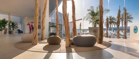 Un vestíbulo moderno con vista al mar, con pilares de madera, zonas de estar y plantas en macetas.