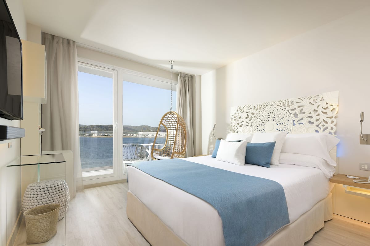 Quarto de hotel moderno com uma cama grande, televisão, mesa de vidro e vista para o mar.