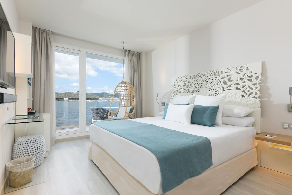 Quarto de hotel moderno com uma cama grande, varanda com vista para o mar, cadeira suspensa e uma pequena escrivaninha com abajur.
