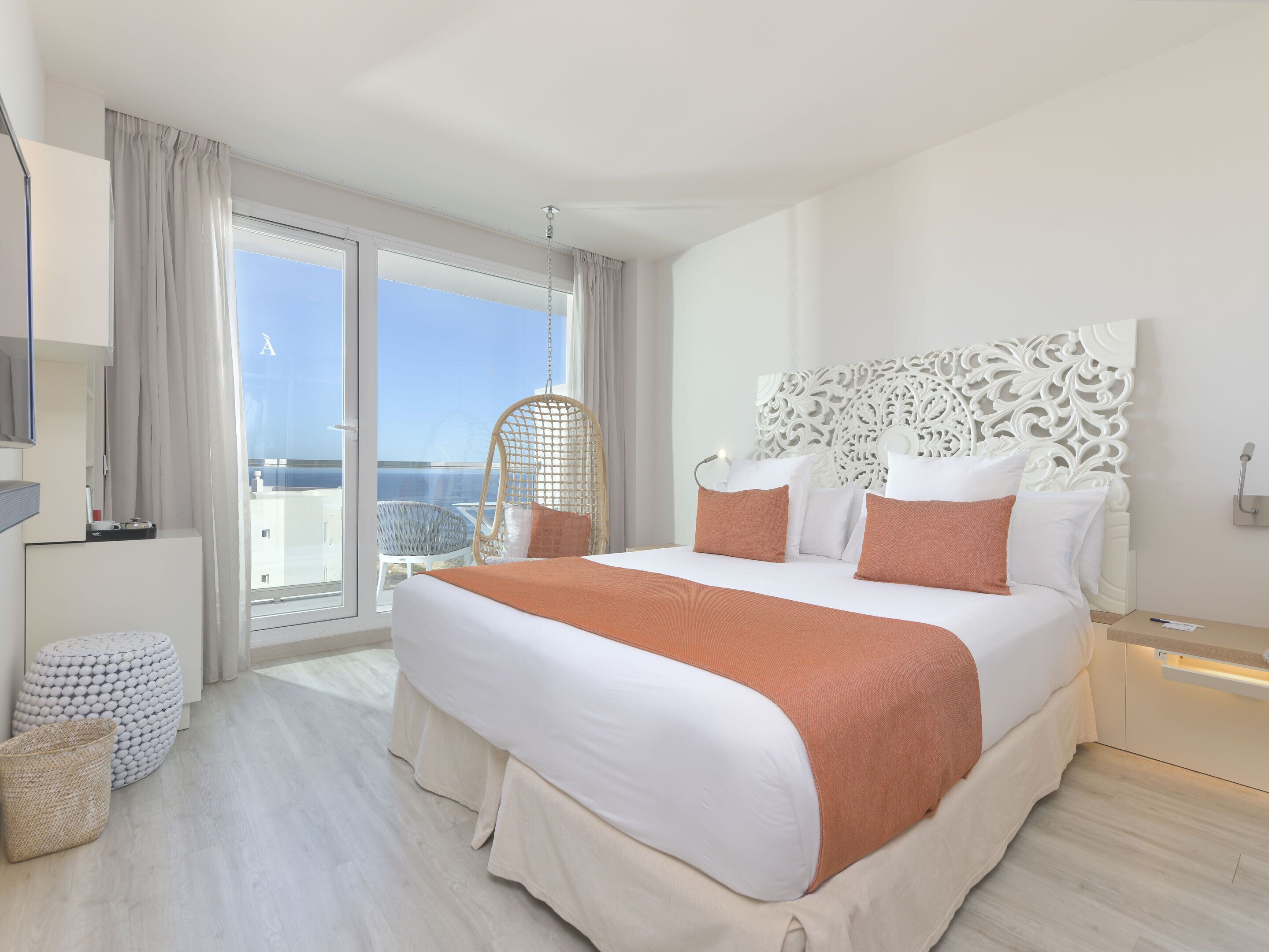 A modern hotel room with a large bed, a hanging chair, and a view of the sea.