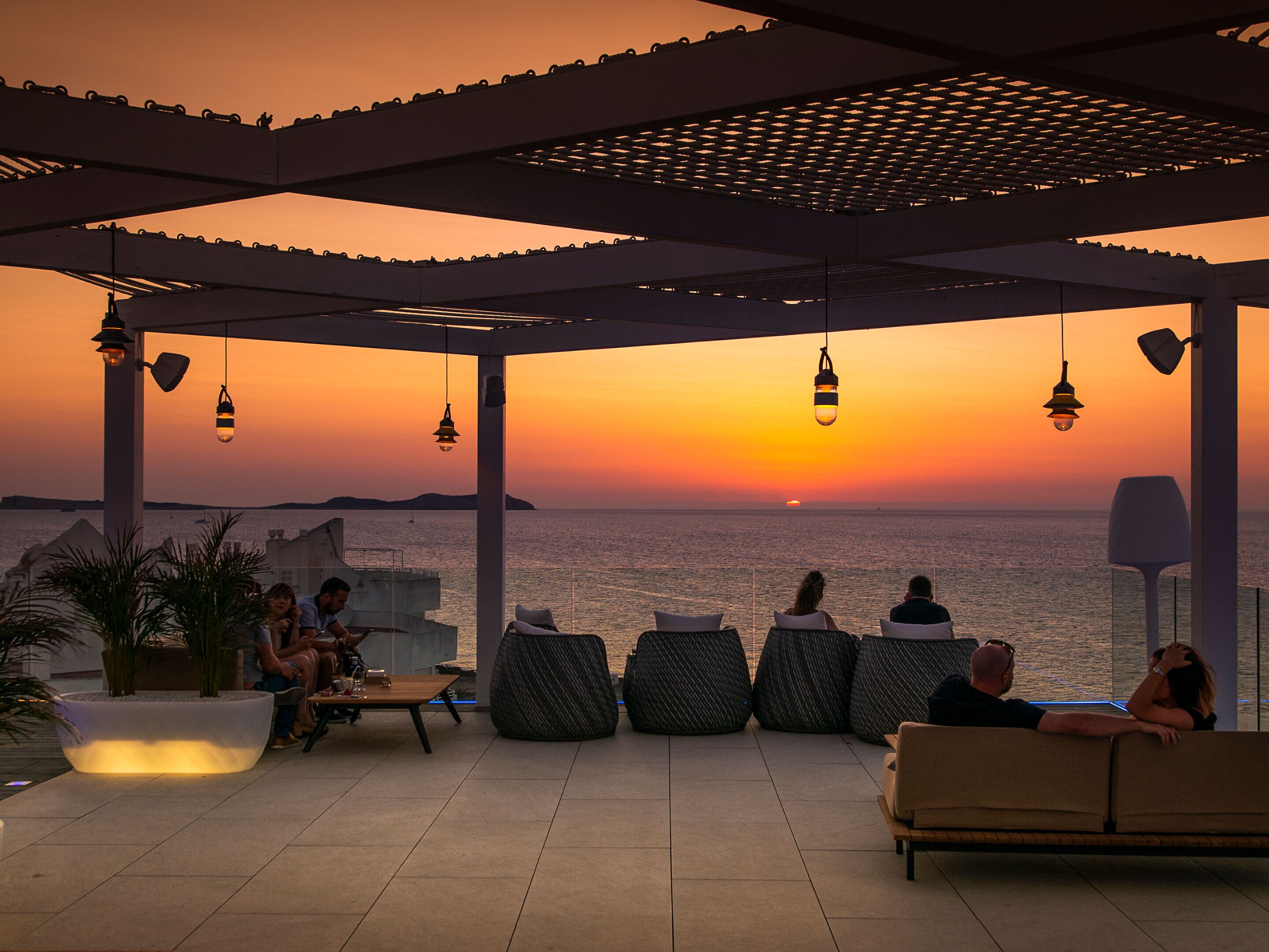 Un espacio al aire libre techado con vistas a la puesta de sol sobre el mar, con sillones cómodos y mesas.