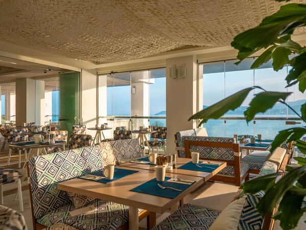 A dining area with patterned furniture, large windows, and a view of the sea. - Amàre Beach Hotel Ibiza - Adults Recommended (Sant Josep de sa Talaia)