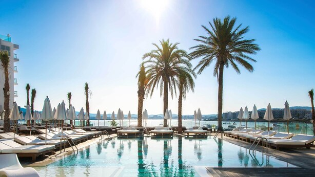 A pool area with lounge chairs and umbrellas, surrounded by palm trees, with a view of the sea and buildings in the background. - Amàre Beach Hotel Ibiza - Adults Recommended (Sant Josep de sa Talaia)