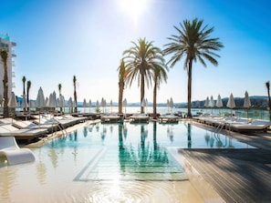 A pool area with lounge chairs and umbrellas, surrounded by palm trees, with a view of the sea and buildings in the background.