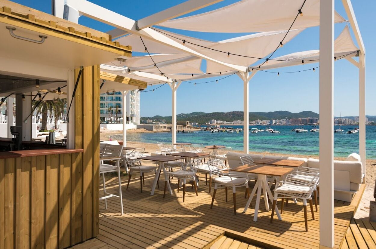 Una terraza al aire libre con mesas y sillas, con vista a una playa y al mar.