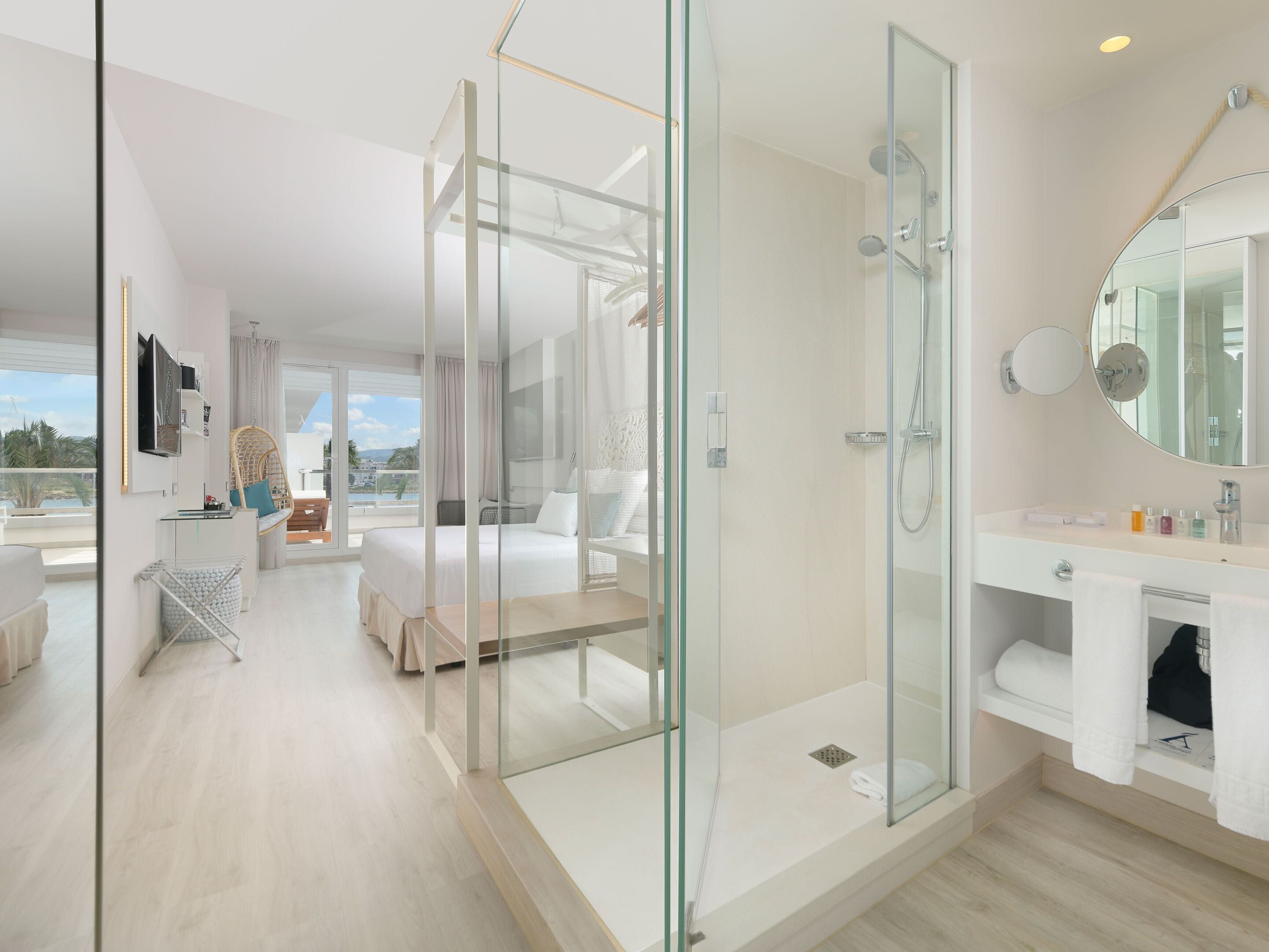 Habitación de hotel moderna con ducha envuelta en vidrio, una cama grande, balcón con vistas y un baño minimalista.